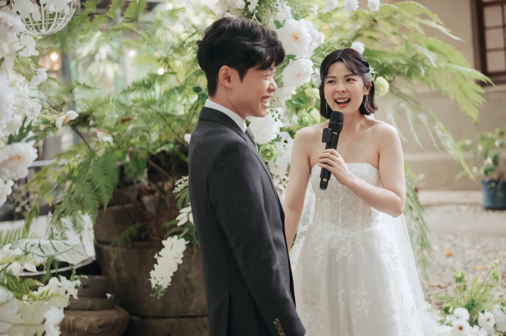 婚禮誓詞紀錄，Tyni 在白色花藝拱門前拿著麥克風與 Farn 分享心意